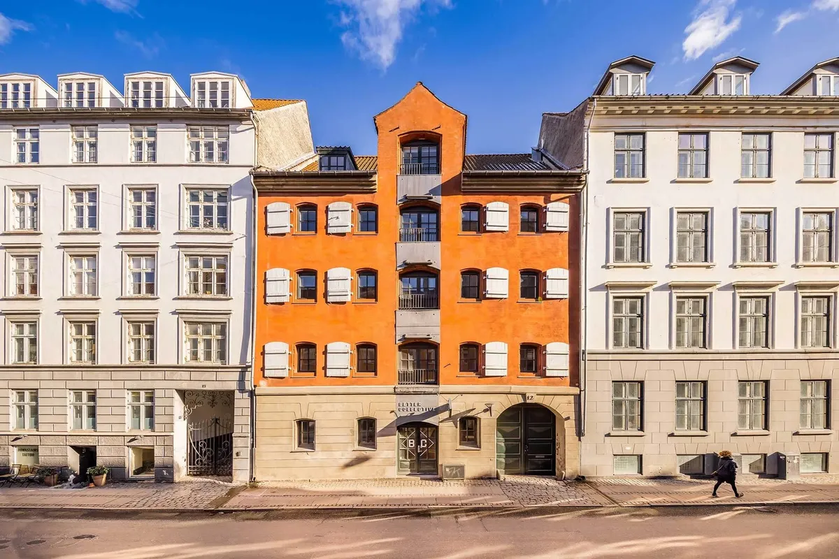 En moderne orange bygning med hvide ruderuder står mellem to hvide bygninger på en brostensbelagt gade i København