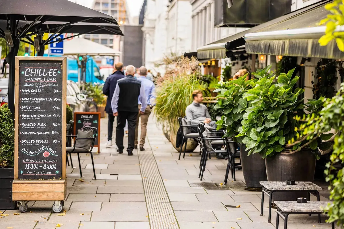 Køkkenhave med spisebord og stole, hvor en mand sidder og nyder lun dag. Der er en menubræt med forskellige sandwich og
