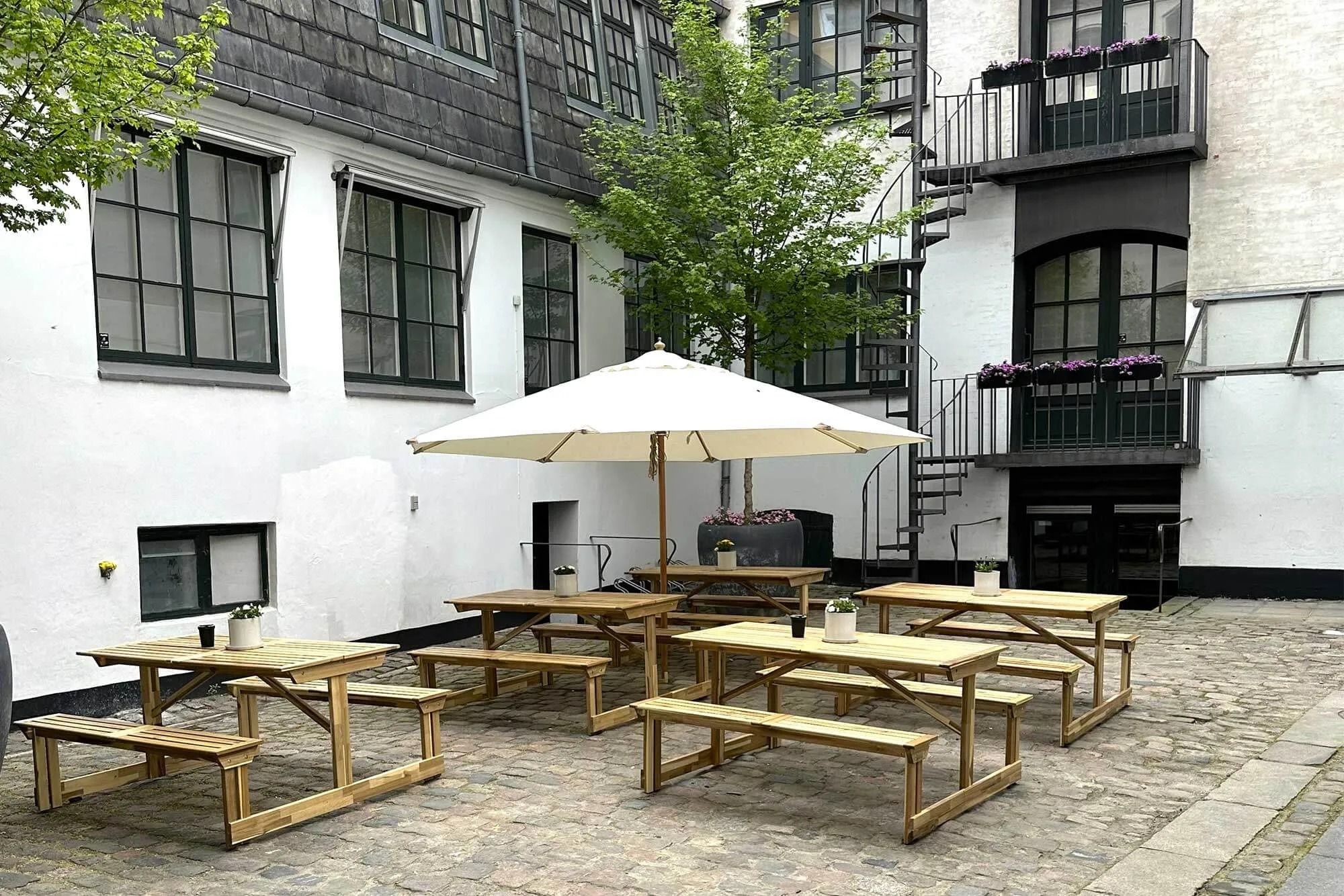 Træborde og bænke udenfor pakhus med skrå tag og vindue med møbelsøjler og planter på altan