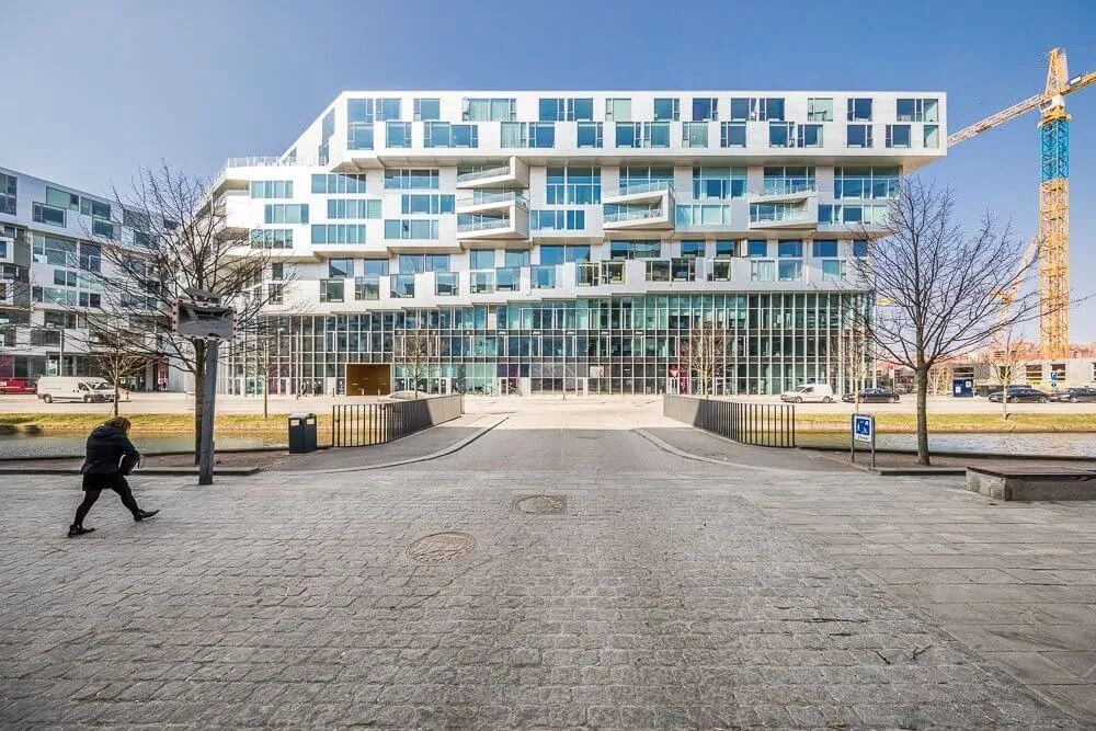 Moderne højhus med glasfacade og balkoner. Person i sort dragt går på brosten. Kran og parkeringsplads med biler. Skov og