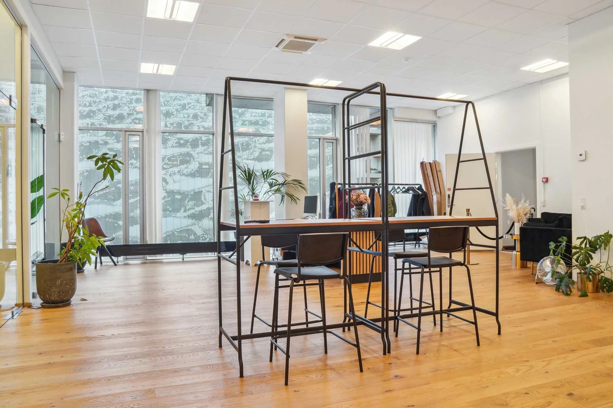 Kontor med møbler og plantager. To stolpeborde med høje stole. Bagved vinduer med udsigt til grønne planter og buske.