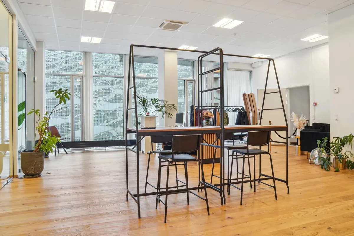 Kontor med møbler og plantager. To stolpeborde med høje stole. Bagved vinduer med udsigt til grønne planter og buske.