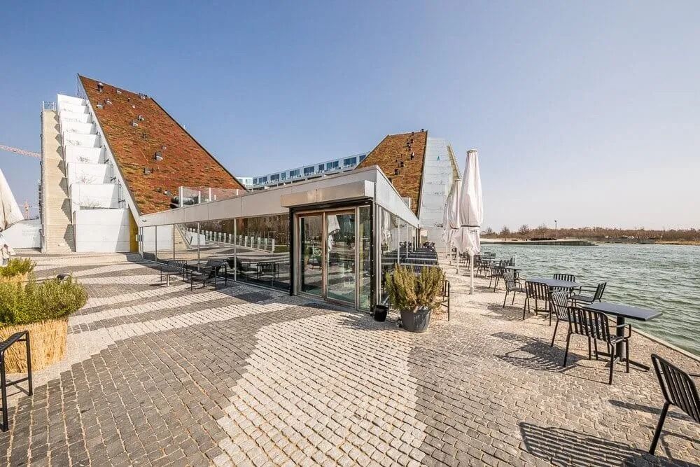 En byggeplads med plantepoteter og møbler samt et restaurantområde med borde og parasoller. Bygningen er tegnet af Bjarke