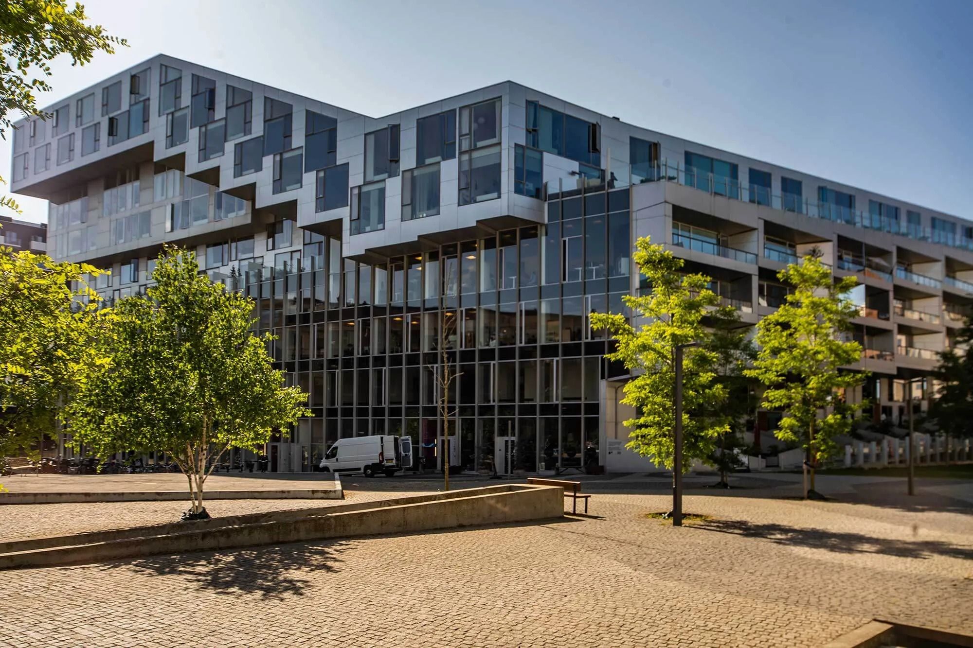 Moderne bygning med glasvinduer og balkoner i København, hvor der er parkerede biler og grønne træer foran