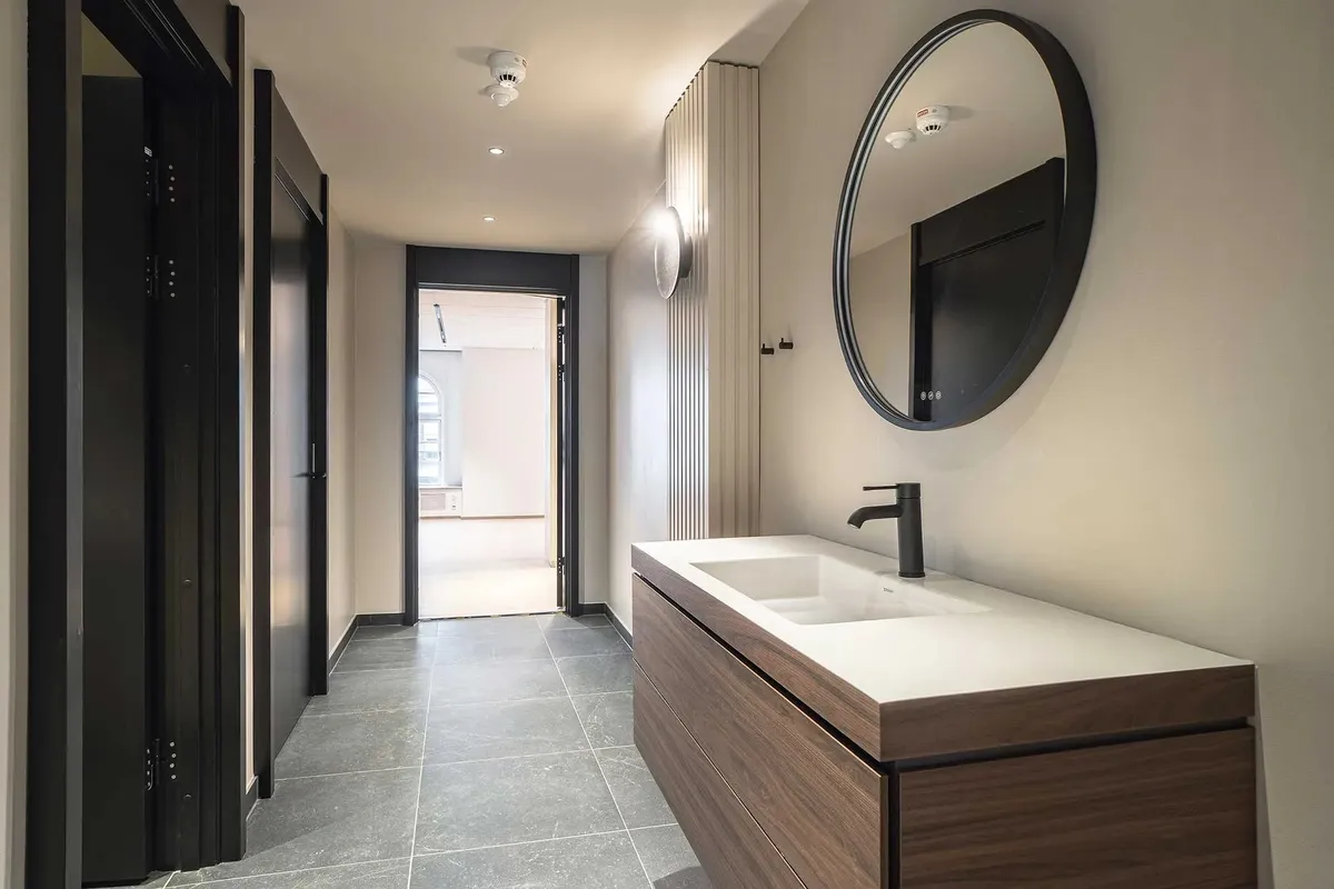 Modernt design med brun træskab, hvid vask og sort spejl med rundt glas. Der er en hvid væg med runde røgdetektorer og en