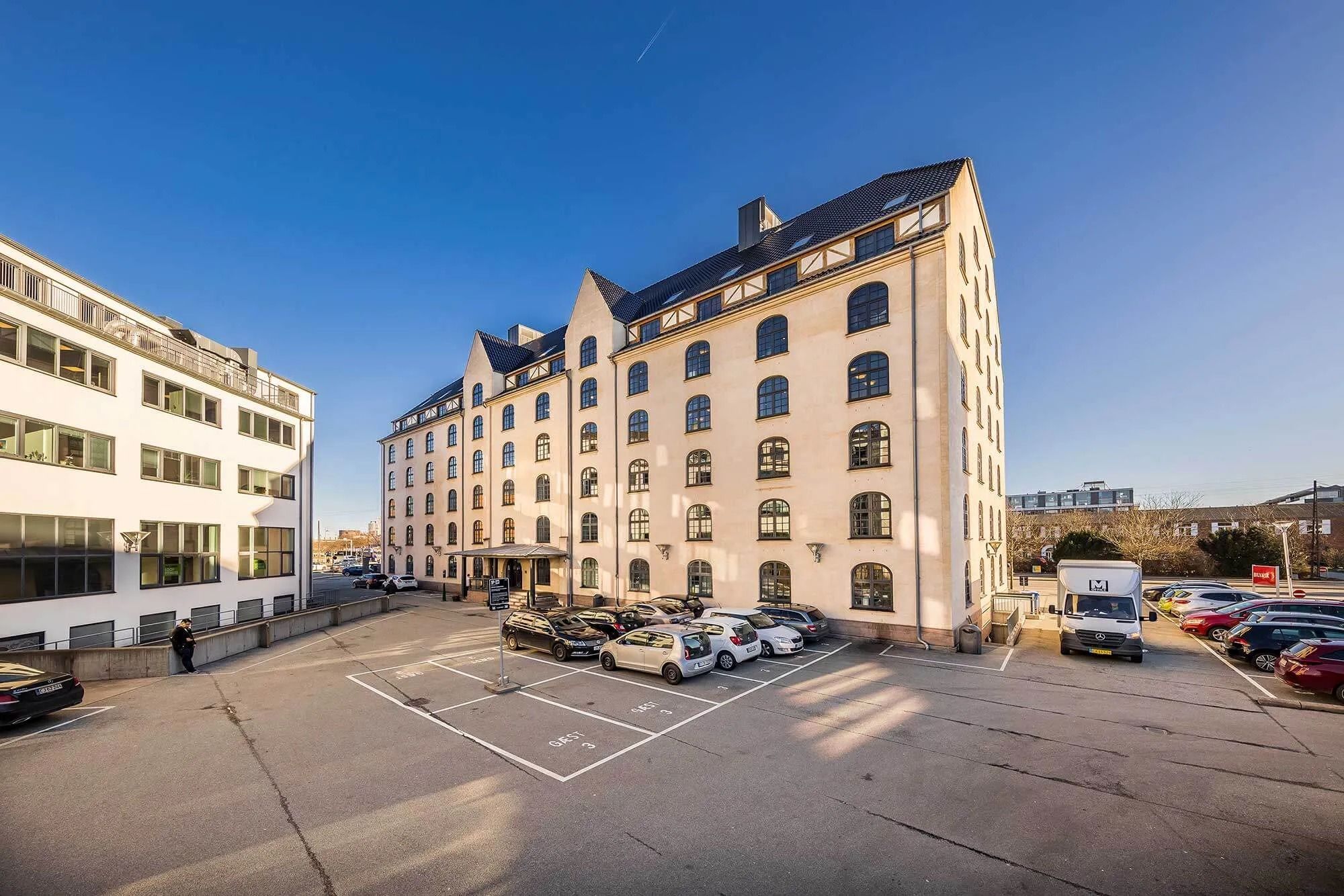 En parkeringsplads med biler parkeret foran et bygningskompleks med flere etager og store vinduer