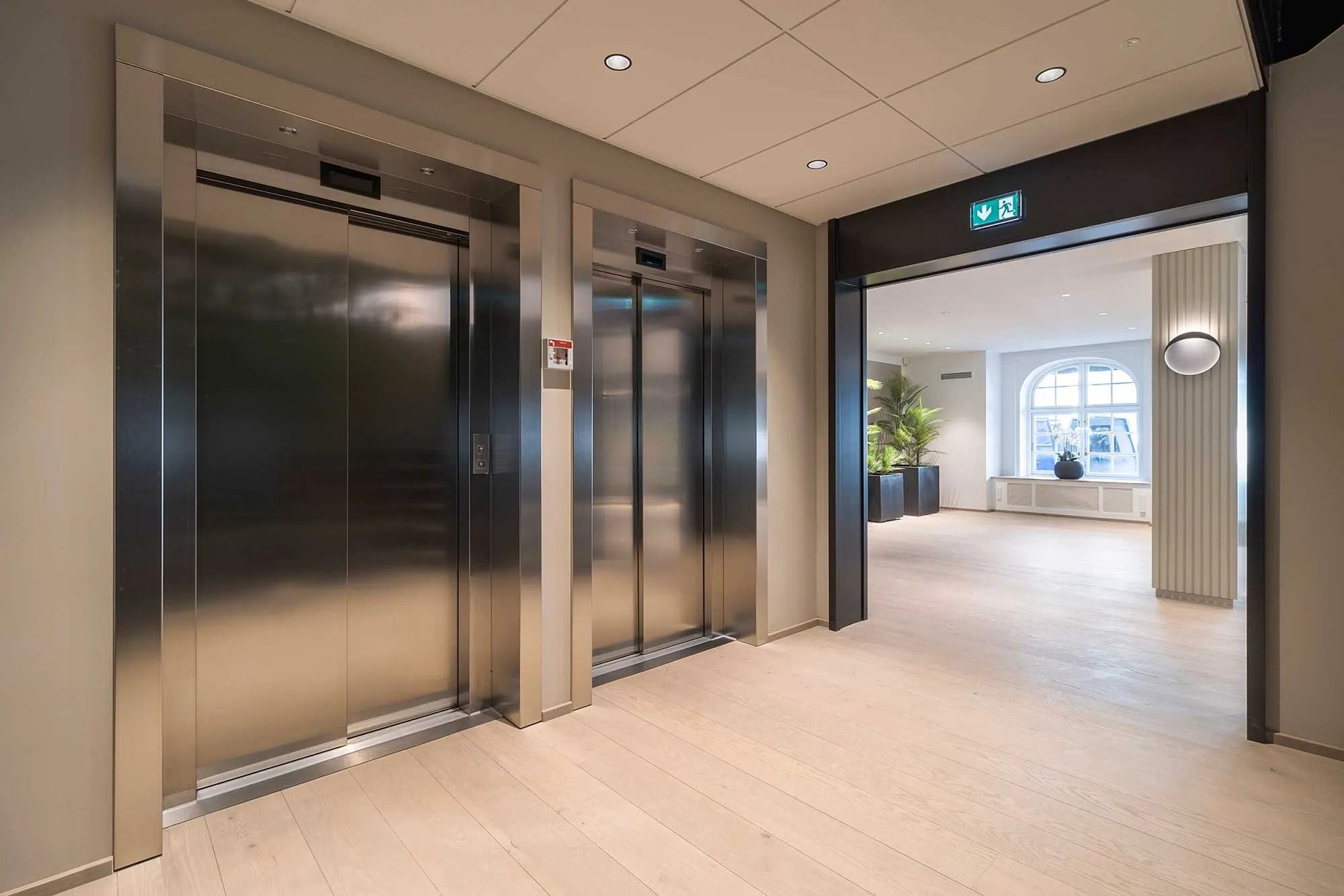To moderne elevatorer står åbne i et kontorlokale med lyse gulve og plantepot. Der er en dør til en anden stue med vinduer