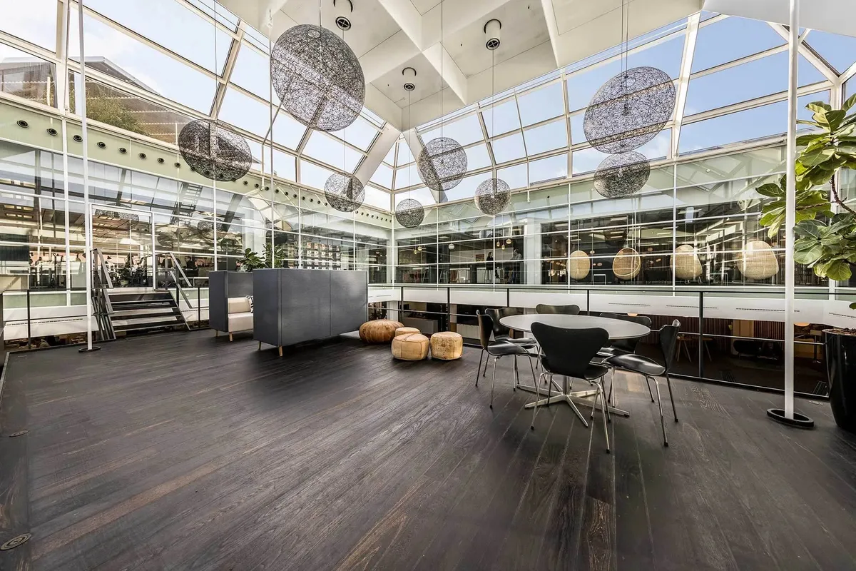 Et kontorlokale med krydsfinergulve, runde stole, bord, sofa og lysekroner samt en stor vinduesfront