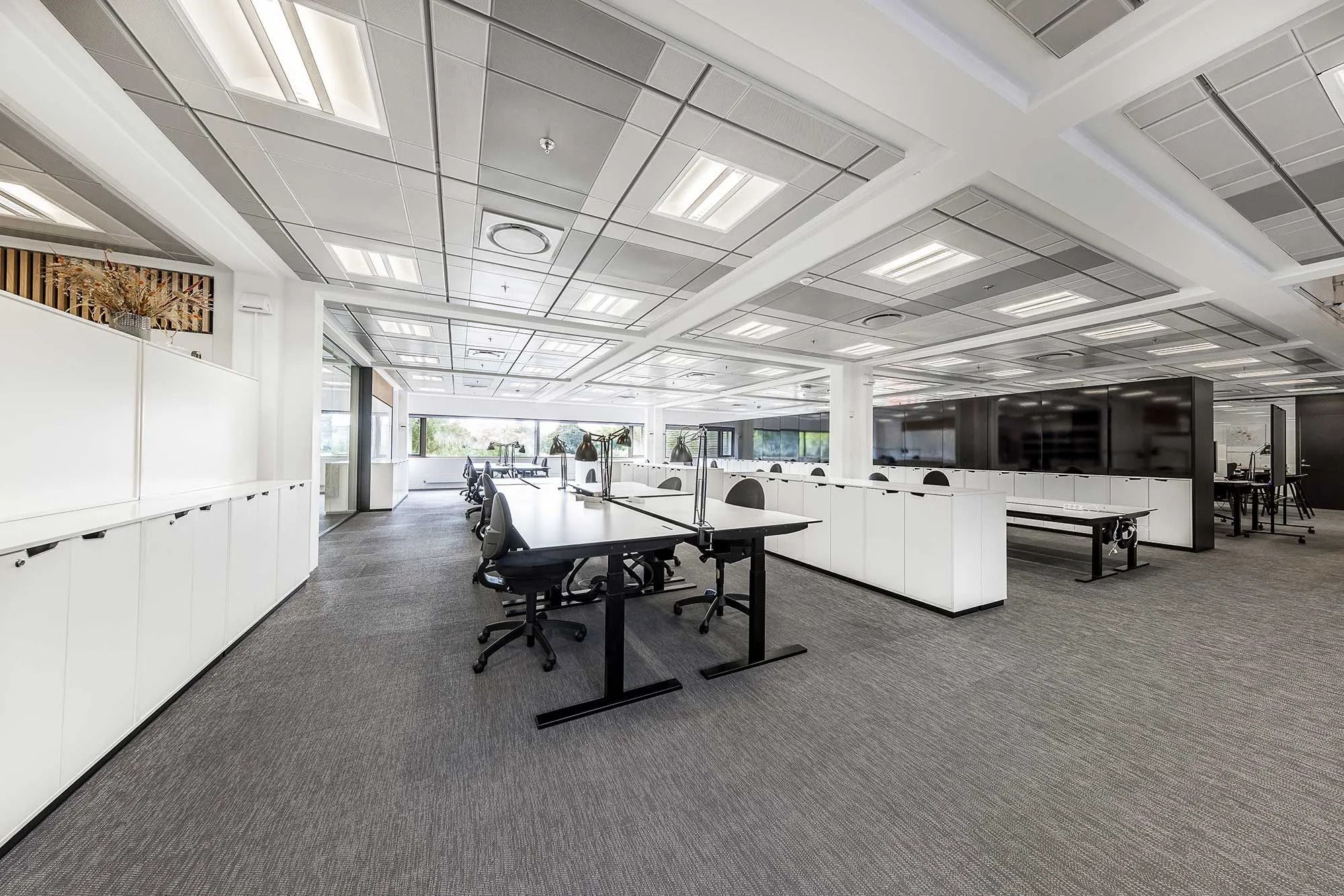 Kontorområde med lyse lofter, hvide vægge, lysegrå gulvbelægning og flere lange borde med sorte stole