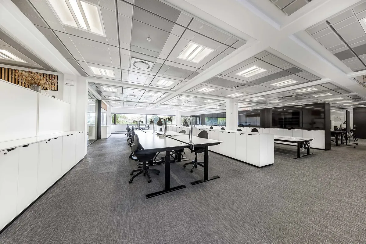 Kontorområde med lyse lofter, hvide vægge, lysegrå gulvbelægning og flere lange borde med sorte stole