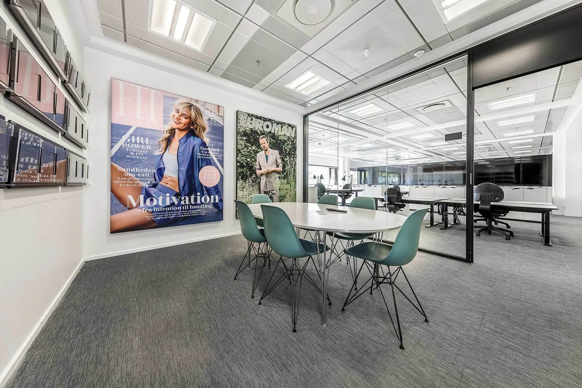 Kontor med bord, stole og glasvægge. På væggen hænger en stor plakat med en kvinde og en mand.