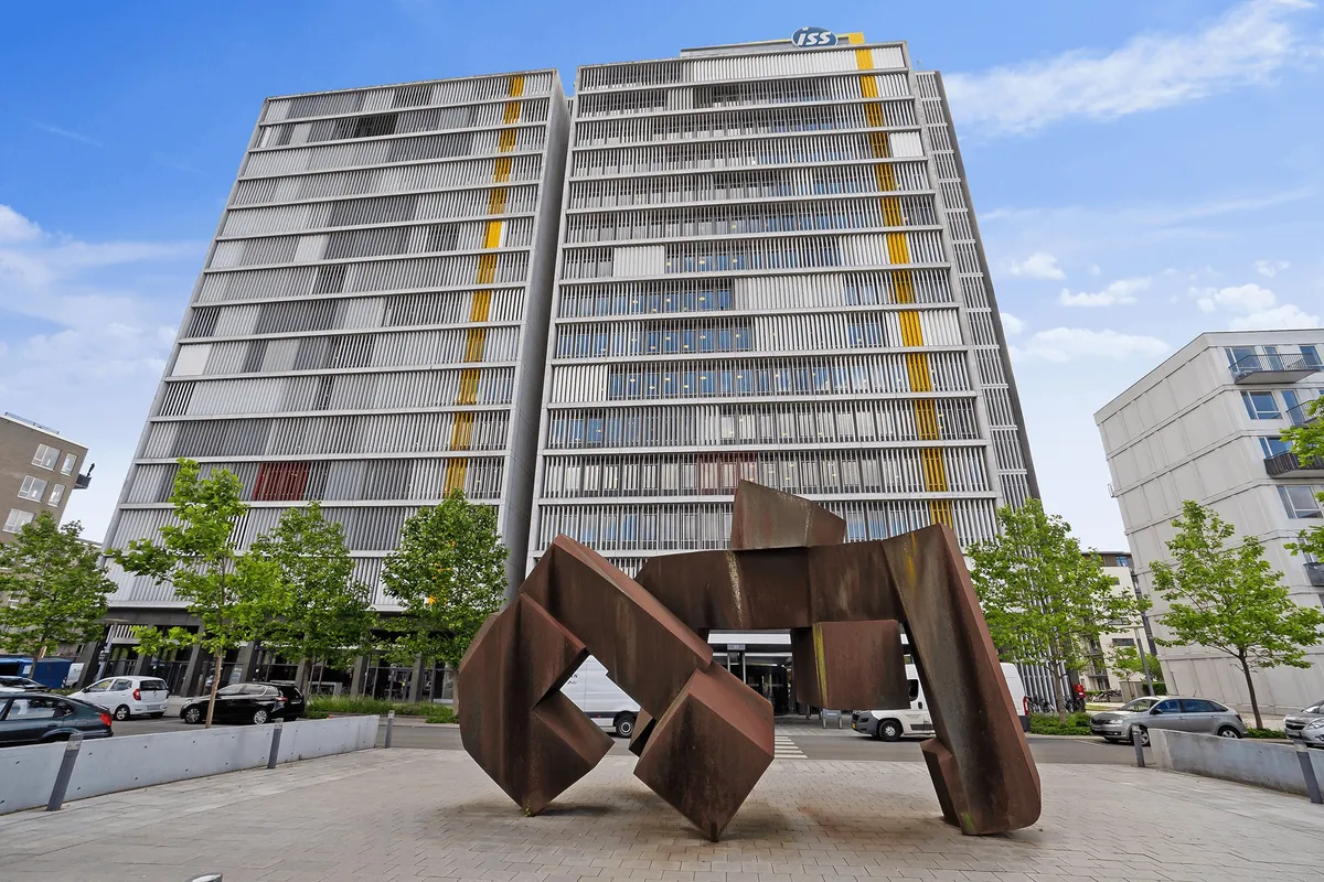 En rusten skulptur står foran et højhus med ISS logoet på toppen. I baggrunden er der en bygning med balkoner og parkerede