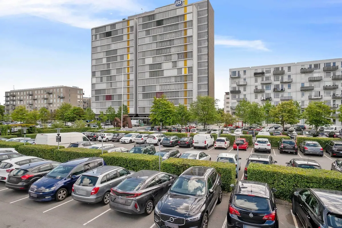 Moderne bygning med lysegul og grå facade placeret midt i en stor parkeringsplads med busser, biler og grønne buske i