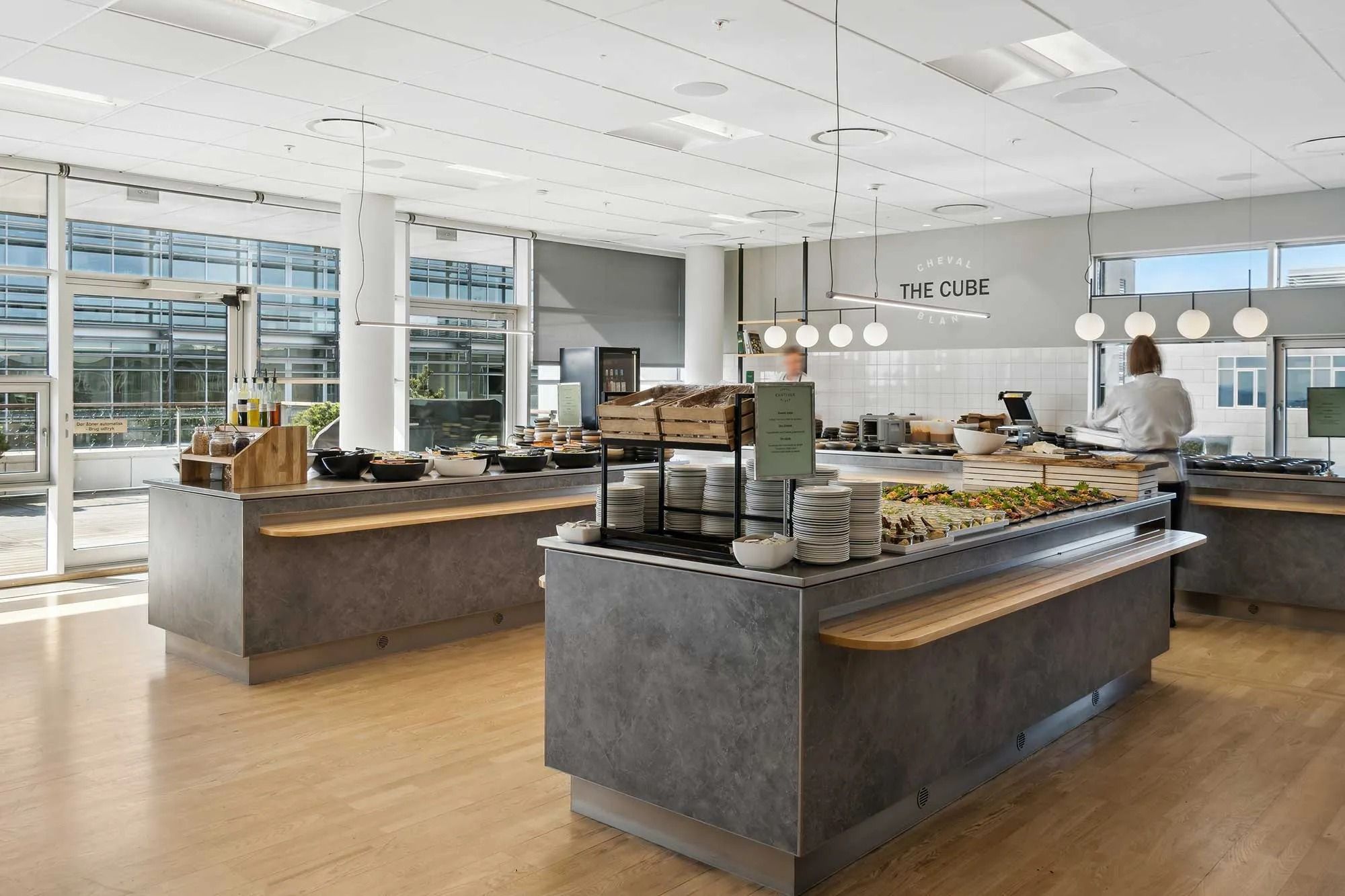 Kanteen med forskellige skåle og tallerkener, krydderier, frugt og grøntsager, og en medarbejder bag disken serverer gæster