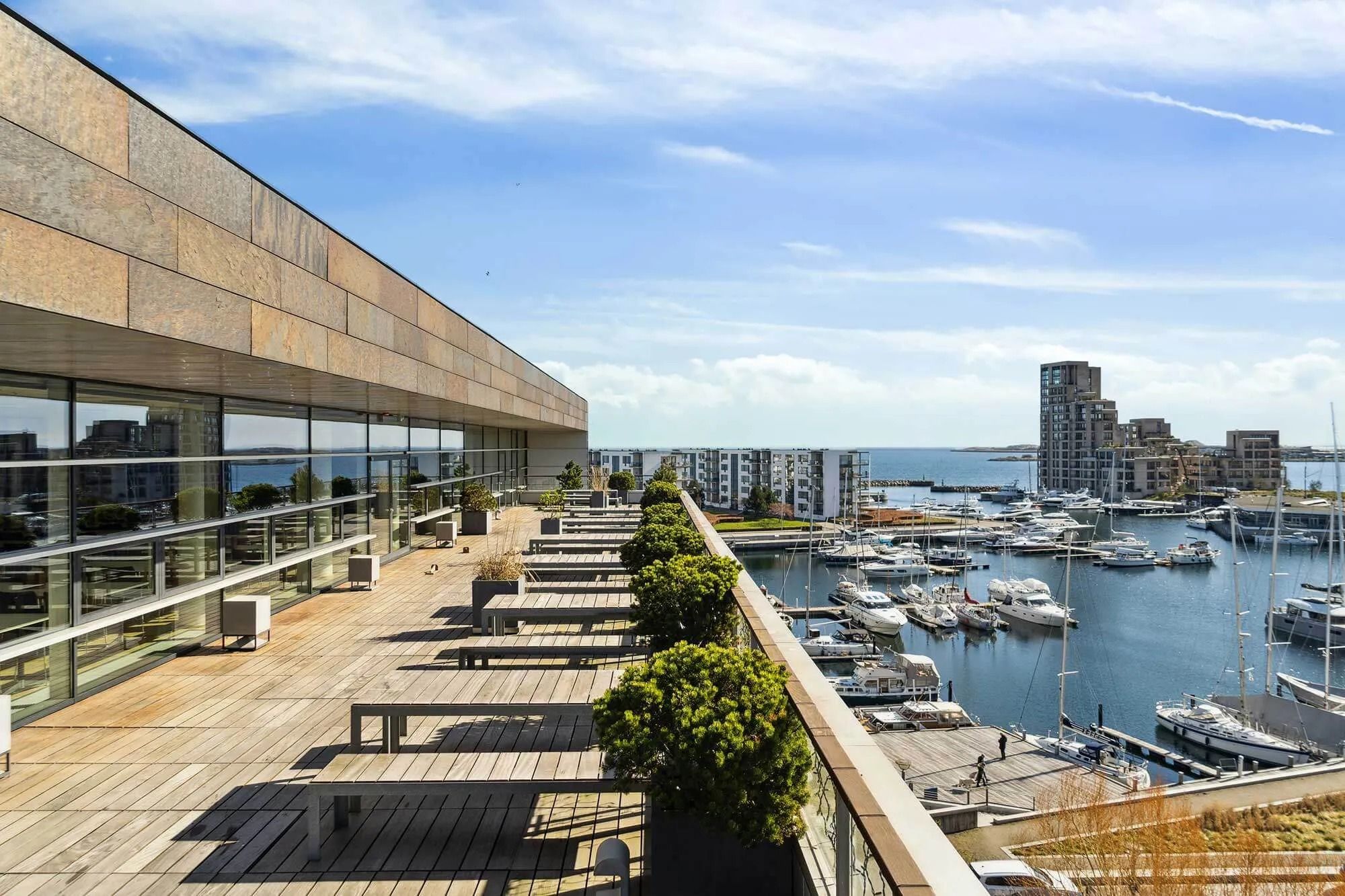 Terrasse med trægulve og udsigt over havn med flere havnefaciliteter og lyse himmel med skyer