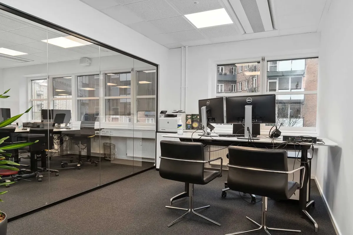 Kontor med glasvæg, høje vinduer, fire Dell-skærme, to sorte stolpeborde, to sort-hvide stolpeborde, fire sort-hvide
