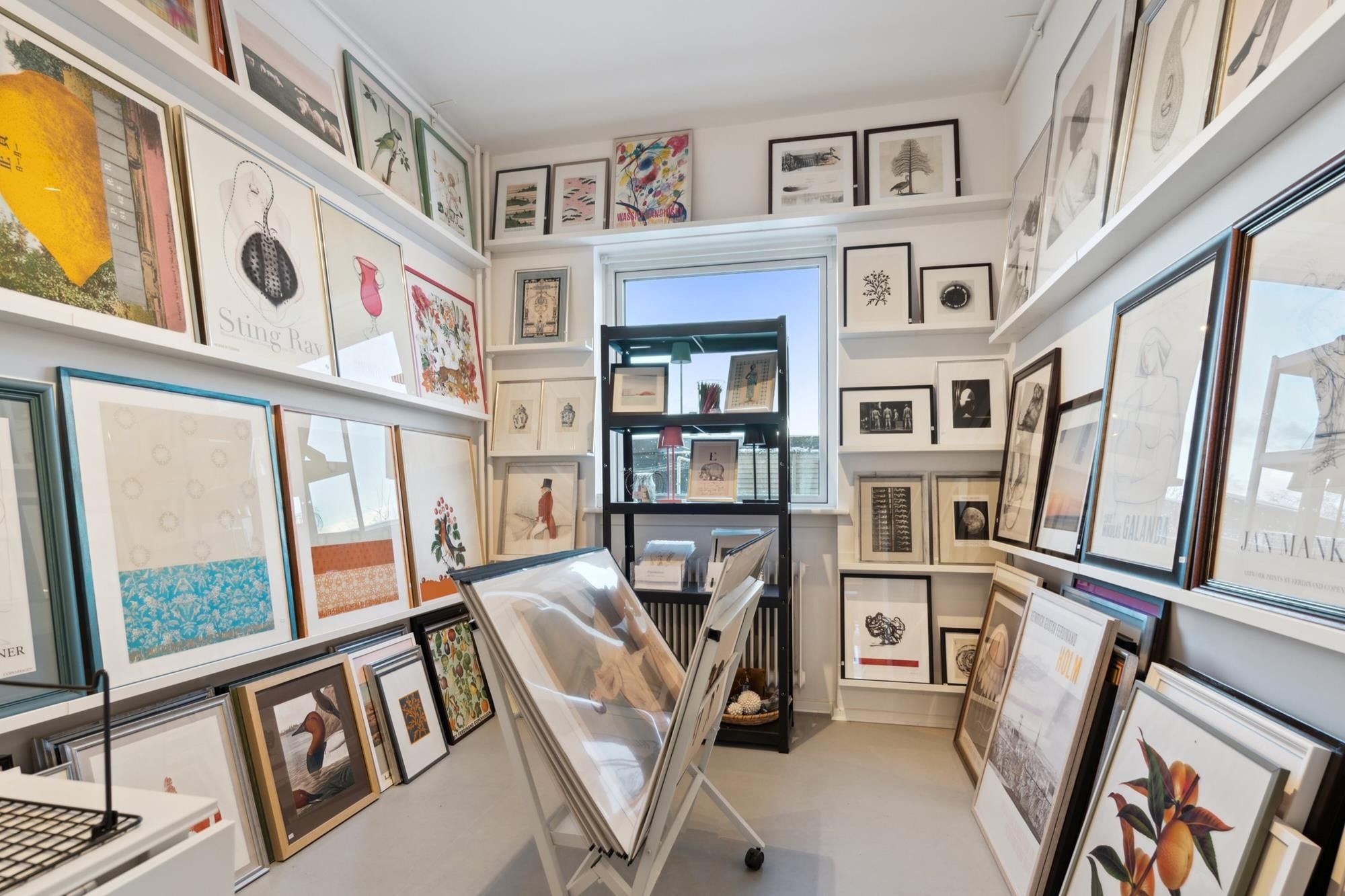 En lille galleri med store hvide hylder fyldt med rammer med malerier og tegninger. Et gulv med høj kvalitet og en åben