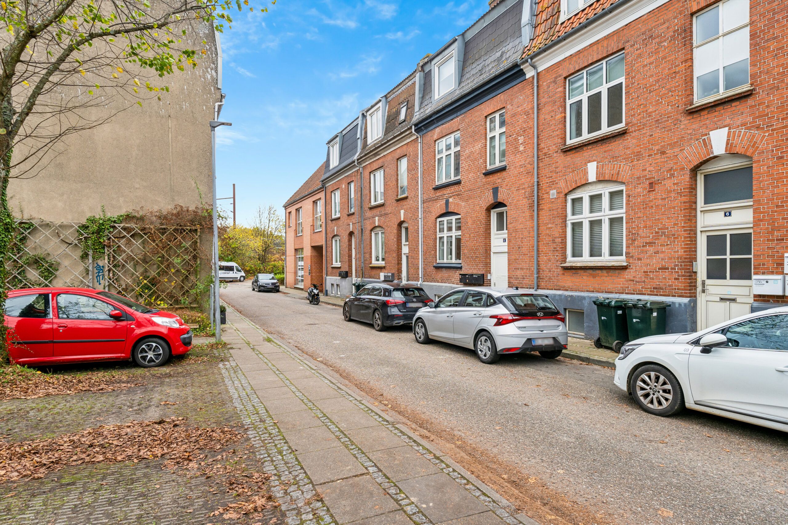 Flere biler parkeret på gade med skur og udsigt til røde og hvide bygninger med mange vinduer og trægrene