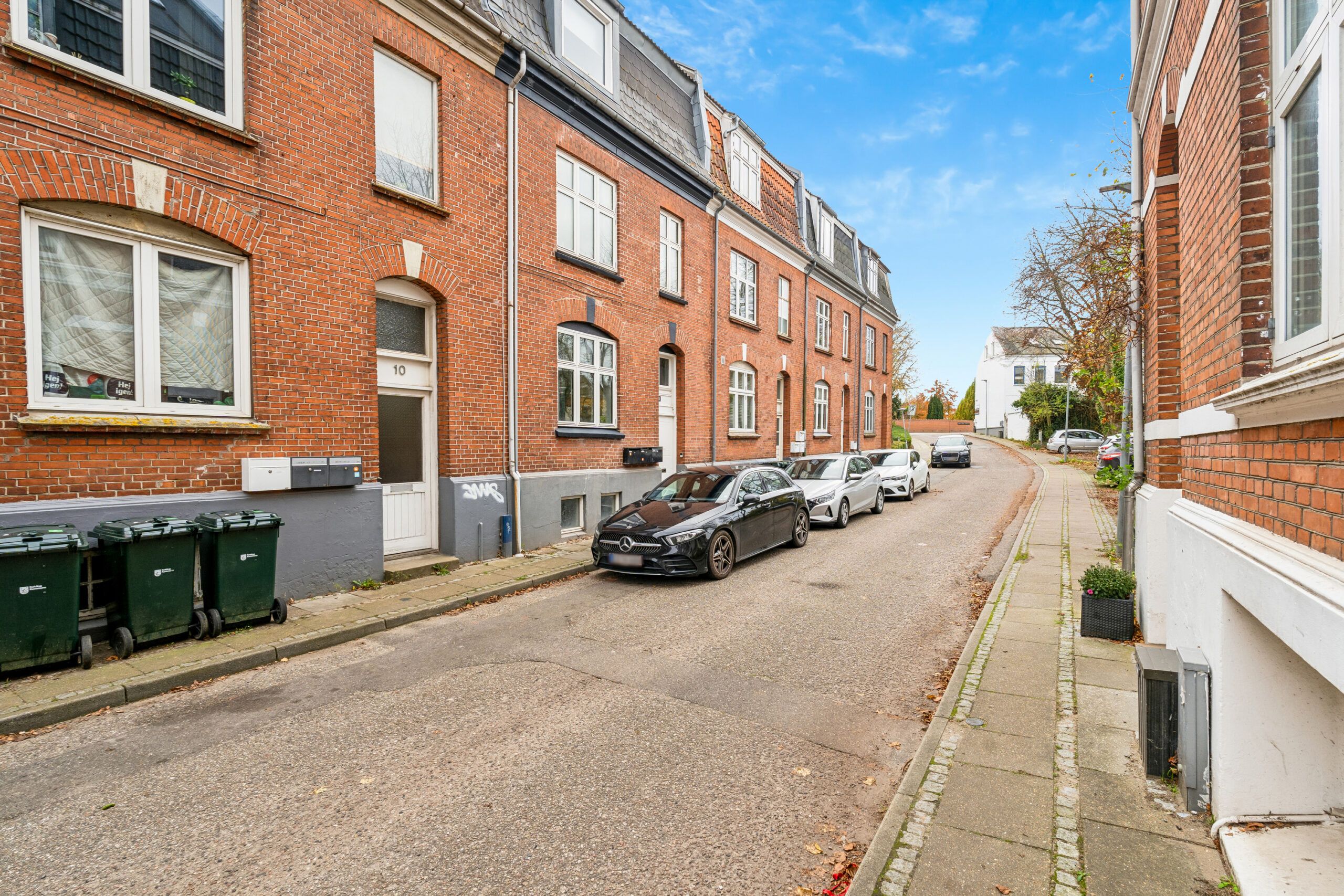 Parkerede biler på en bred asfaltvej med gavlmuren på en side. En Mercedes med hvide forlygter står bag en bil. Et par