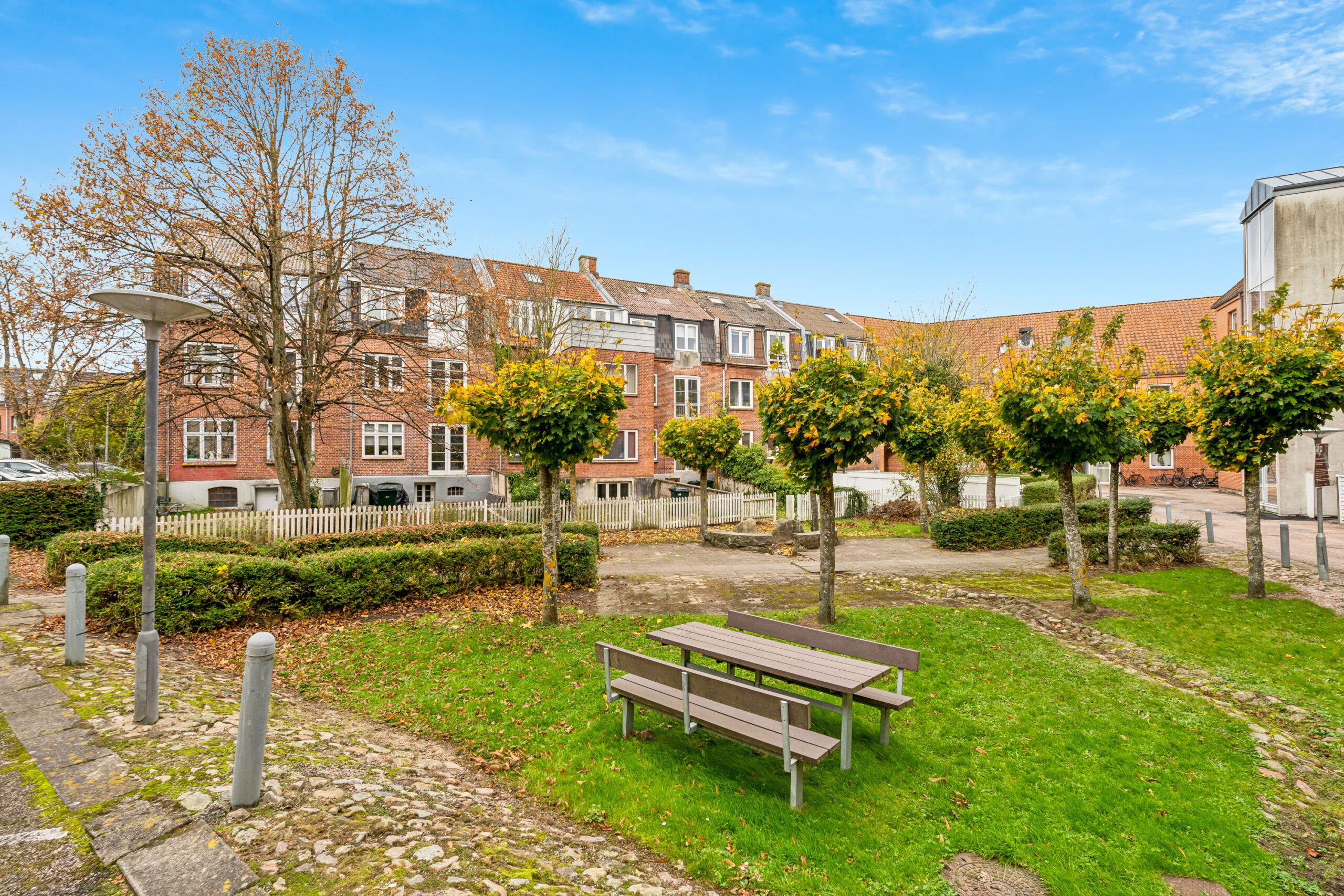 Moderne park med grønne træer, buske og 2 bænke med et rundt bord, ligesom et hus med røde mursten og hvide ruder i