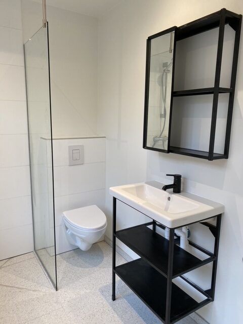 Minimalistisk badeværelse med hvid gulv, hvid toilet og brusekabine med glasdør og håndbruser samt højt stående sort hylde