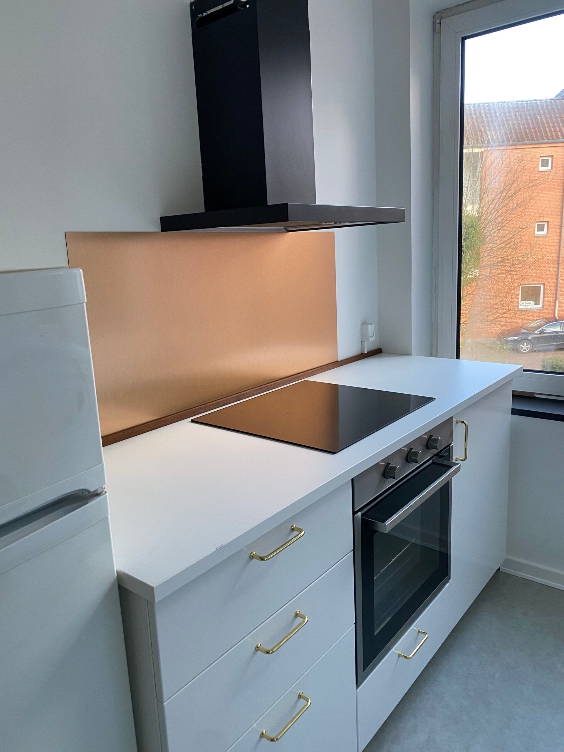 Moderne køkken med lysegrå vægge og hvide skabe med guldfarvede håndtag, kogeplade og ovn med glasdør, køleskab og