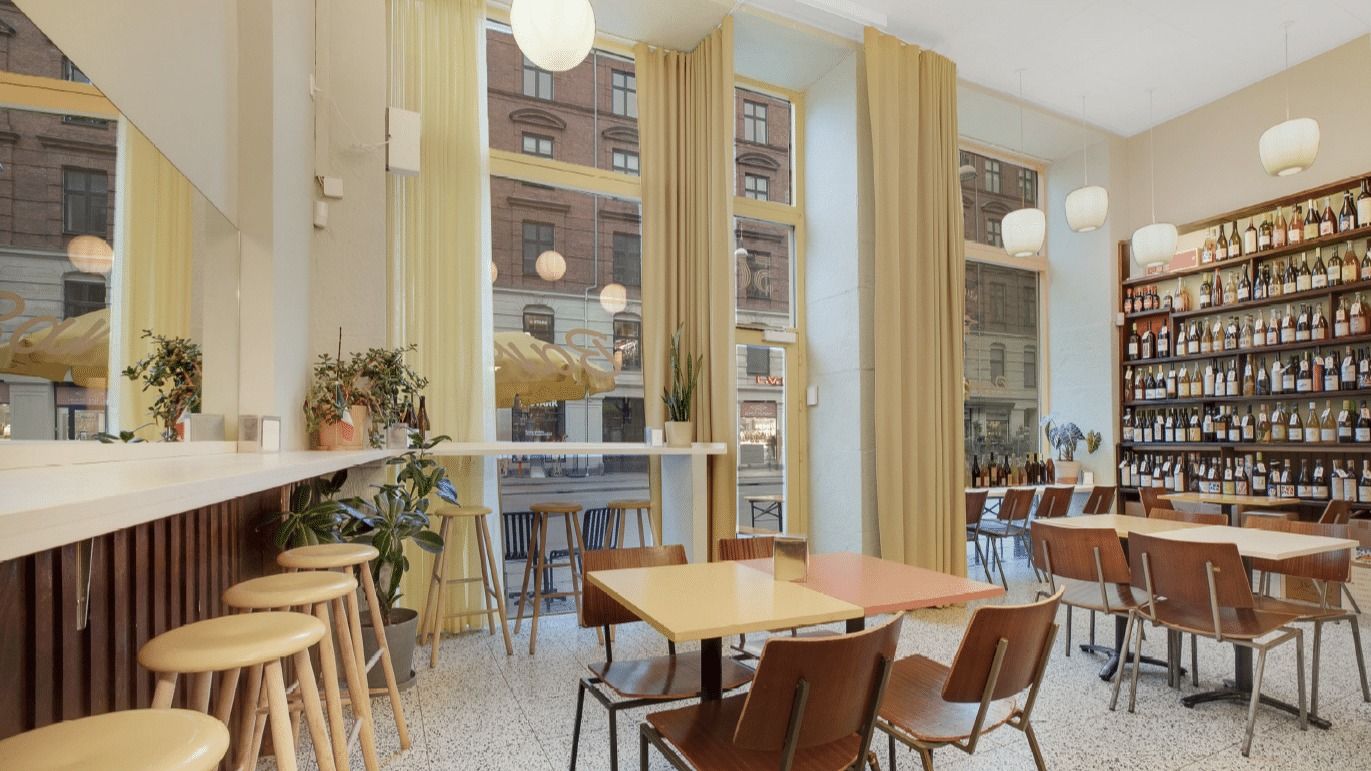 En hyggeligt indrettet café med lysegulle vægge og loftslamper med lysereflekser. Der er flere bord og stole samt høje