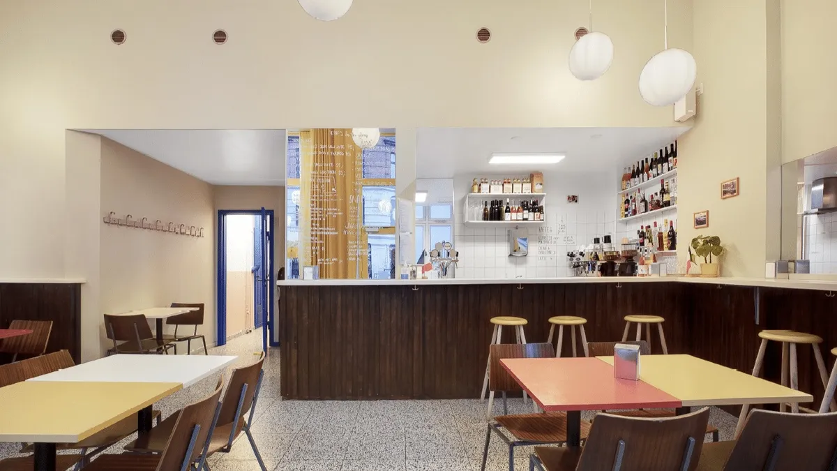 Interiør med højt loft, lyse loftslamper, lyserøde og gule bord og stole og baren i baggrunden med en række flasker og