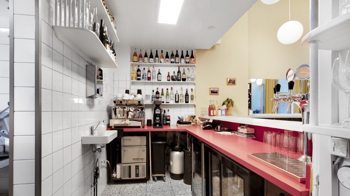 En indret kaffebar med moderne design og røde borde. Væggene er hvide og lyse. Et skab med mange flasker og et sæt glas.