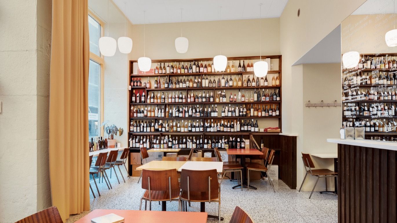 Hyggeligt vinbar med masser af vinflasker på hylder og træborde og stole med lyse loftslamper