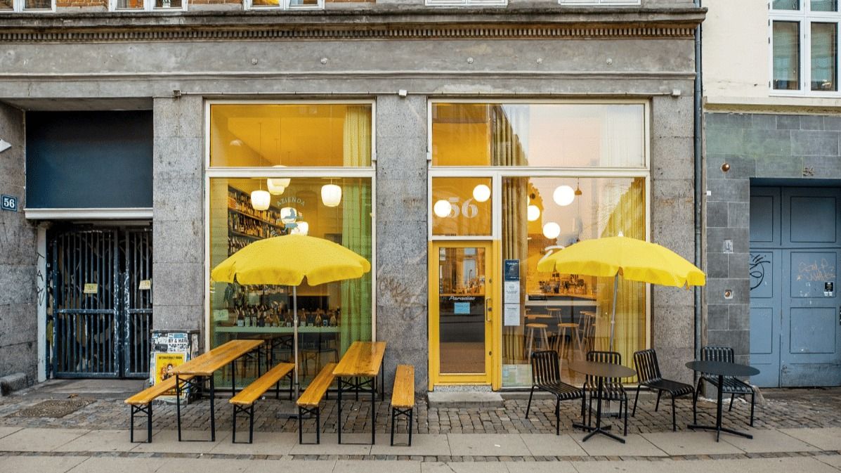 Lyse gule parasoller med bord og stole udenfor et lokalt butiksvindue i København
