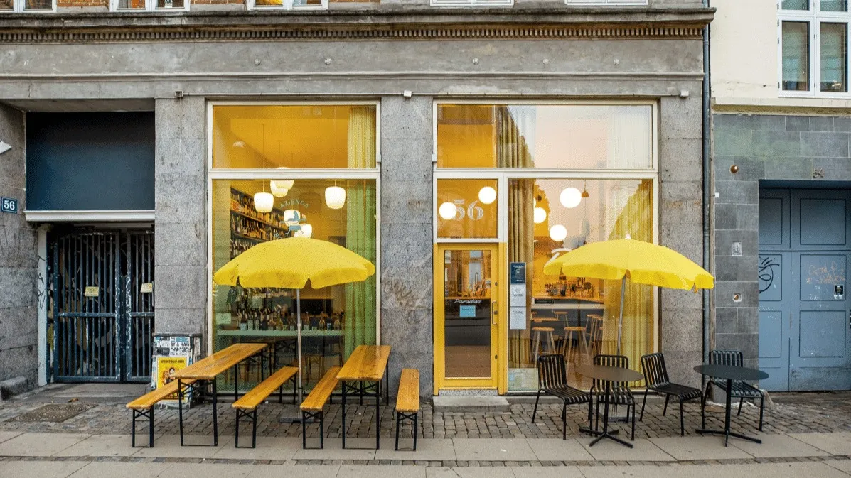 Lyse gule parasoller med bord og stole udenfor et lokalt butiksvindue i København
