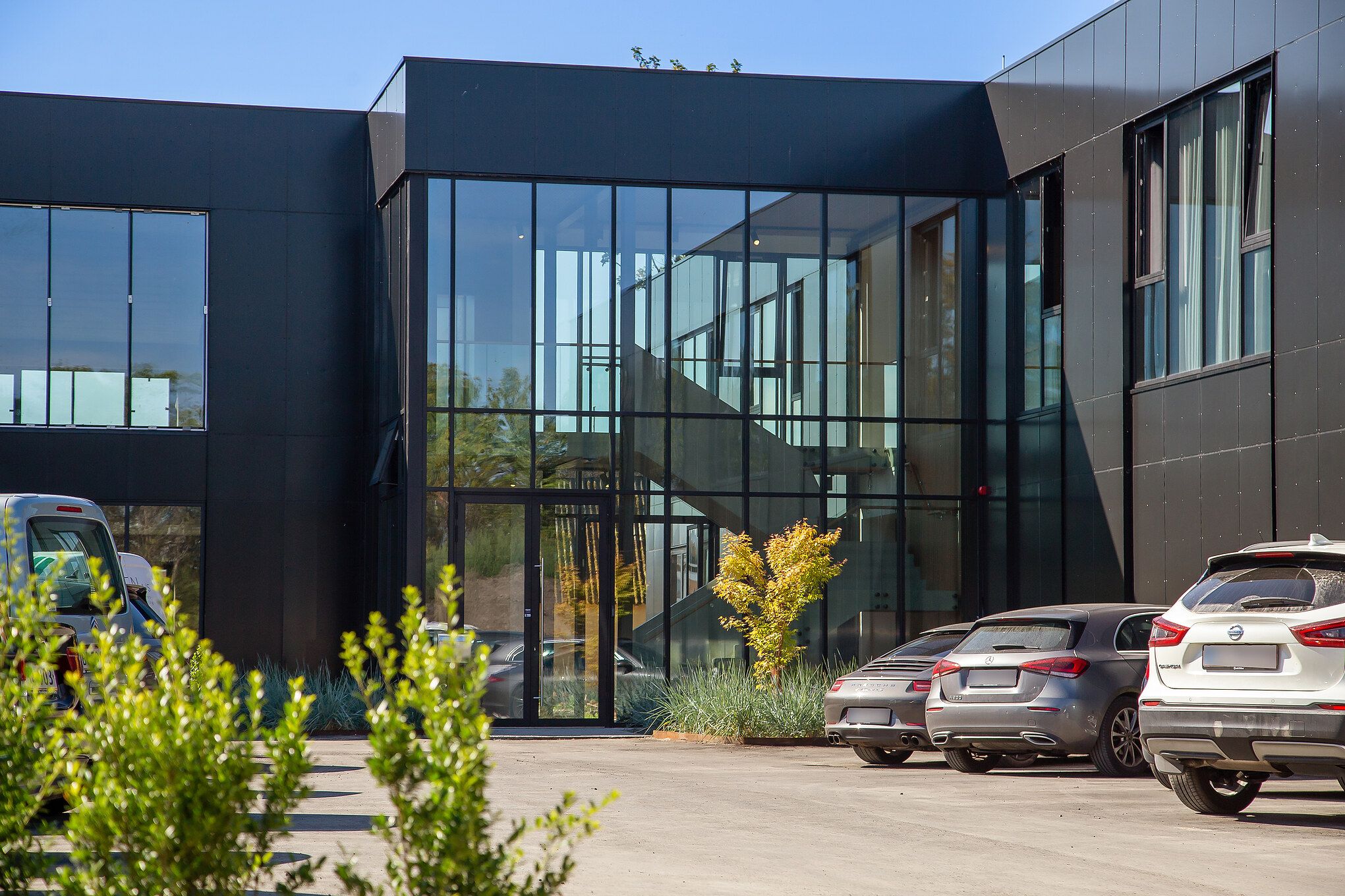 Foran en moderne bygning er der flere biler parkeret, der er et stort glasdør og et glasvindue med spejle, ligesom der er
