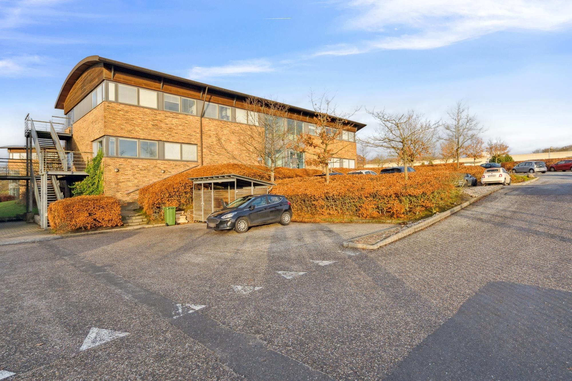 Moderne kontorejendom med grønne buske og flere parkerede biler foran hovedindgangen og trapper til første etage