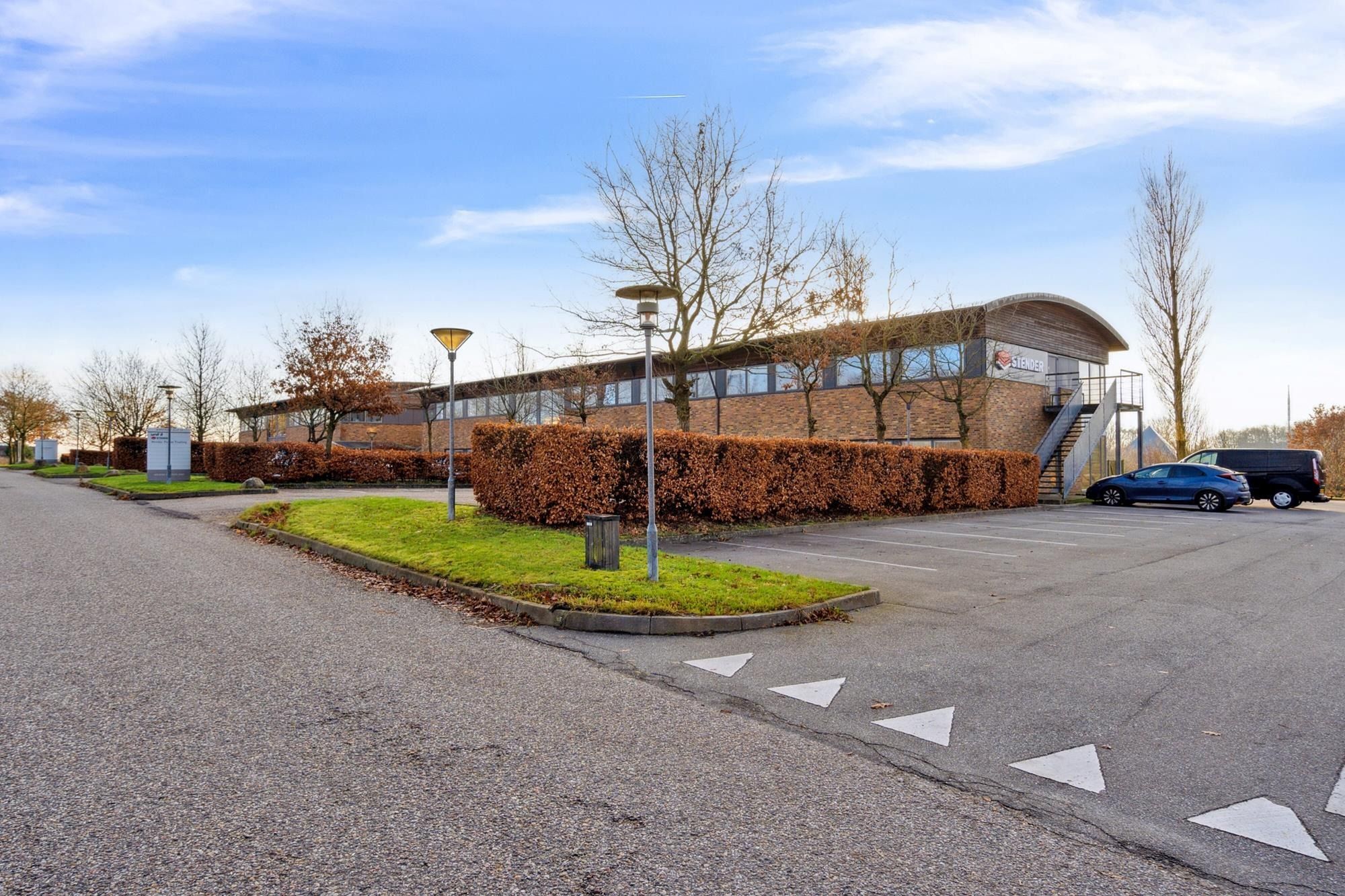 Moderne bygning med glasruder og skilte, hvor Stender er skrevet. En parkeringsplads med to biler parkeret. En række træer