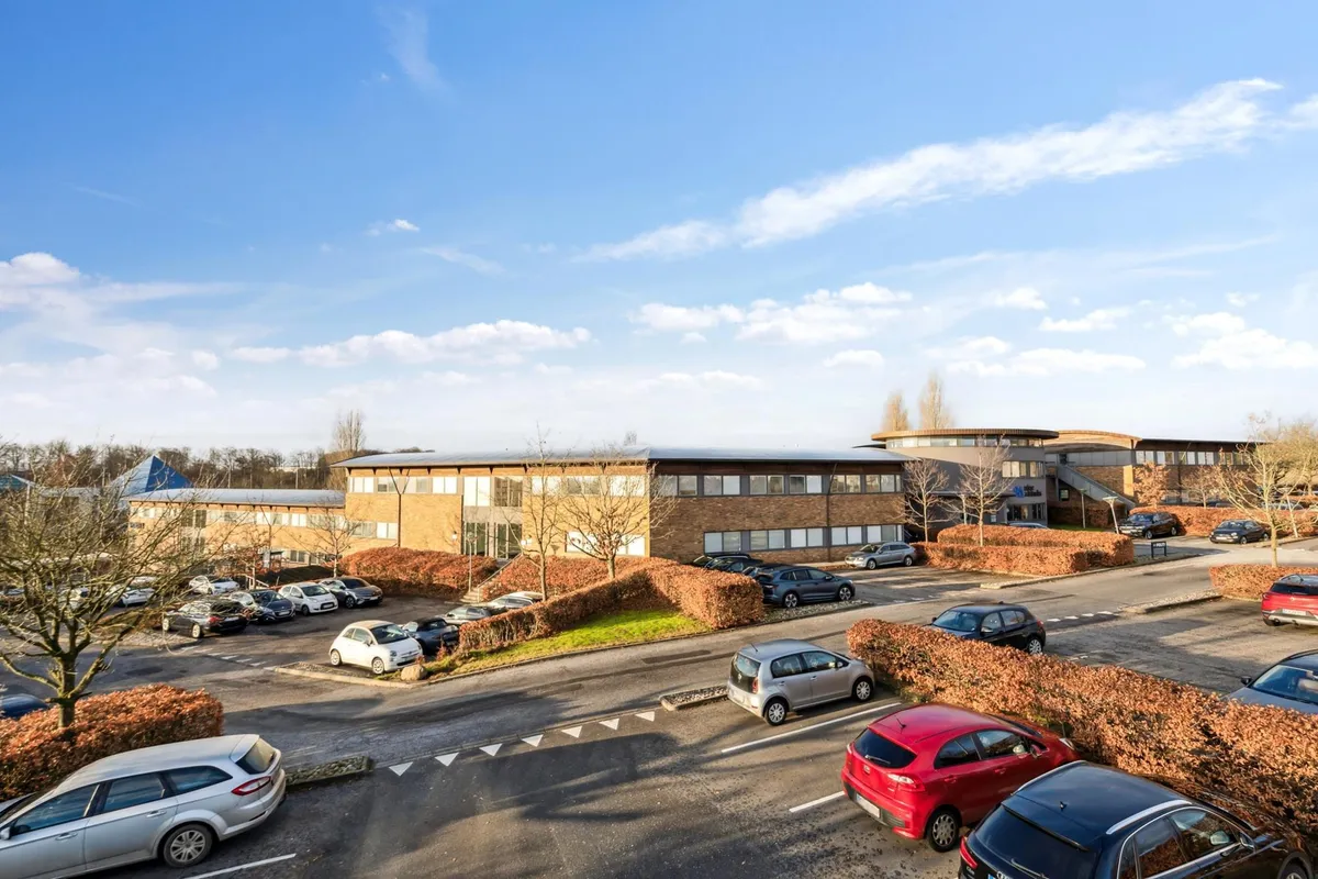 Moderne bygning med parkeringsplads og flere biler parkeret foran og omkring bygningen i solrigt og blåt vejr