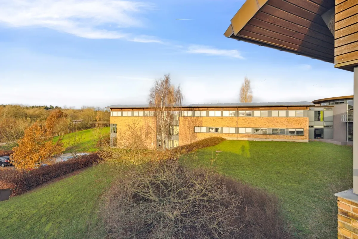 Moderne kontorejendom med lyse vinduerne og terrassen, omgivet af grønne områder og parkeringsplads