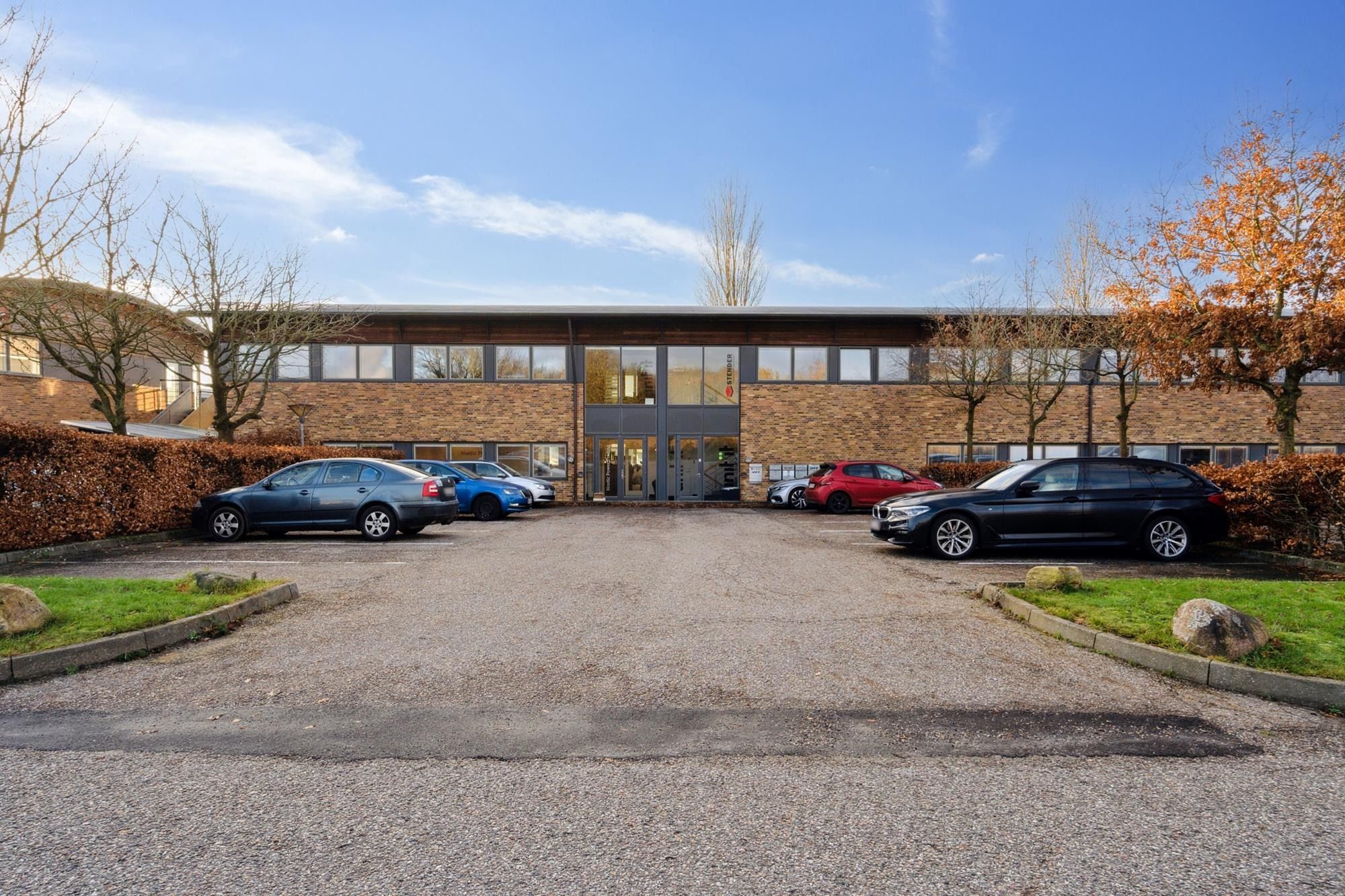 Moderne kontorejendom med glasvinduer og lyse interiører samt parkeringsplads med biler parkeret og smukke træer omkring