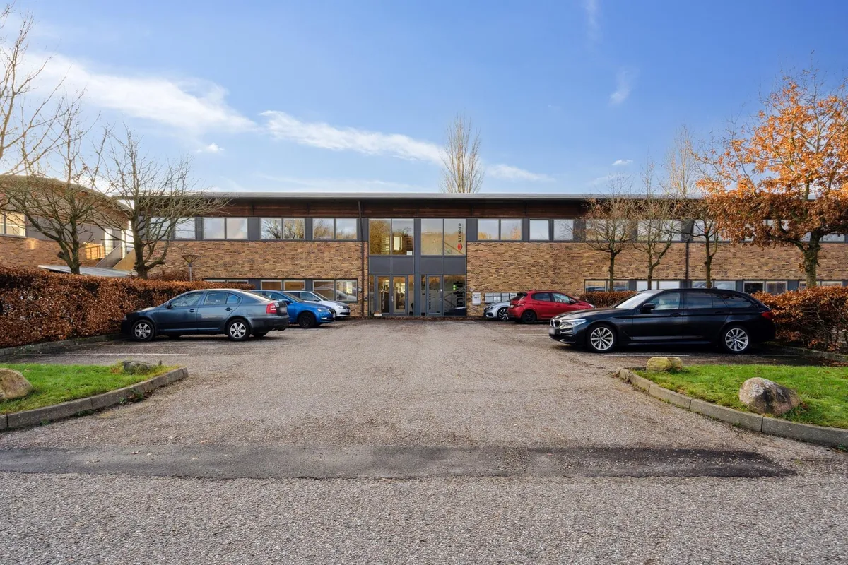 Moderne kontorejendom med glasvinduer og lyse interiører samt parkeringsplads med biler parkeret og smukke træer omkring