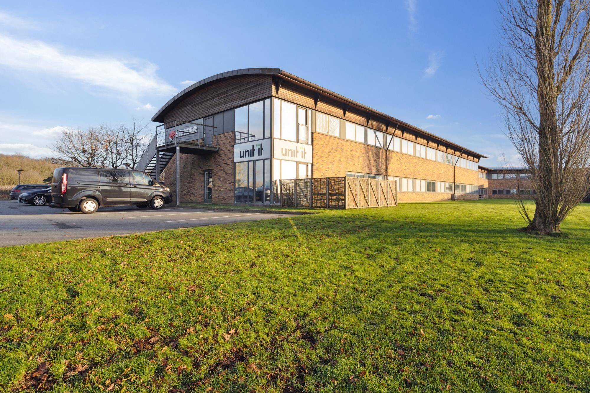 Kontorbyggeri med moderne design, glasruder og entre med skrånende tag i lysebrune mursten. Parkplads med stationeret bil og