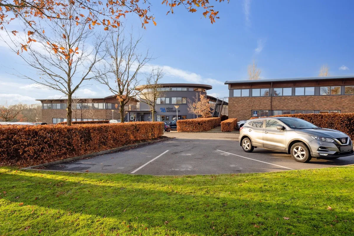 Nissan Qashqai parket på parkeringsplads foran moderne kontorejendom med tæt buskhave og sammenhængende bygninger med