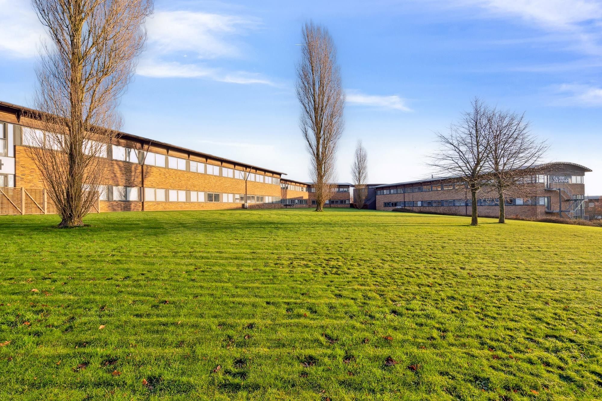 Bredt grønt græsområde med træer og to bygninger med glasvinduer og terrasser på en solrig dag