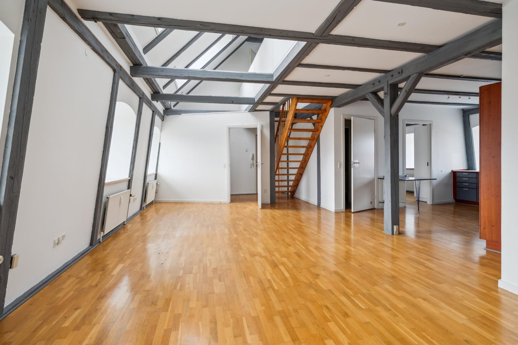 Et stort loftsrum med højloft og masser af lys. En trætrappe og flere døre er synlige. Gulvet er lavet af træ og ser både