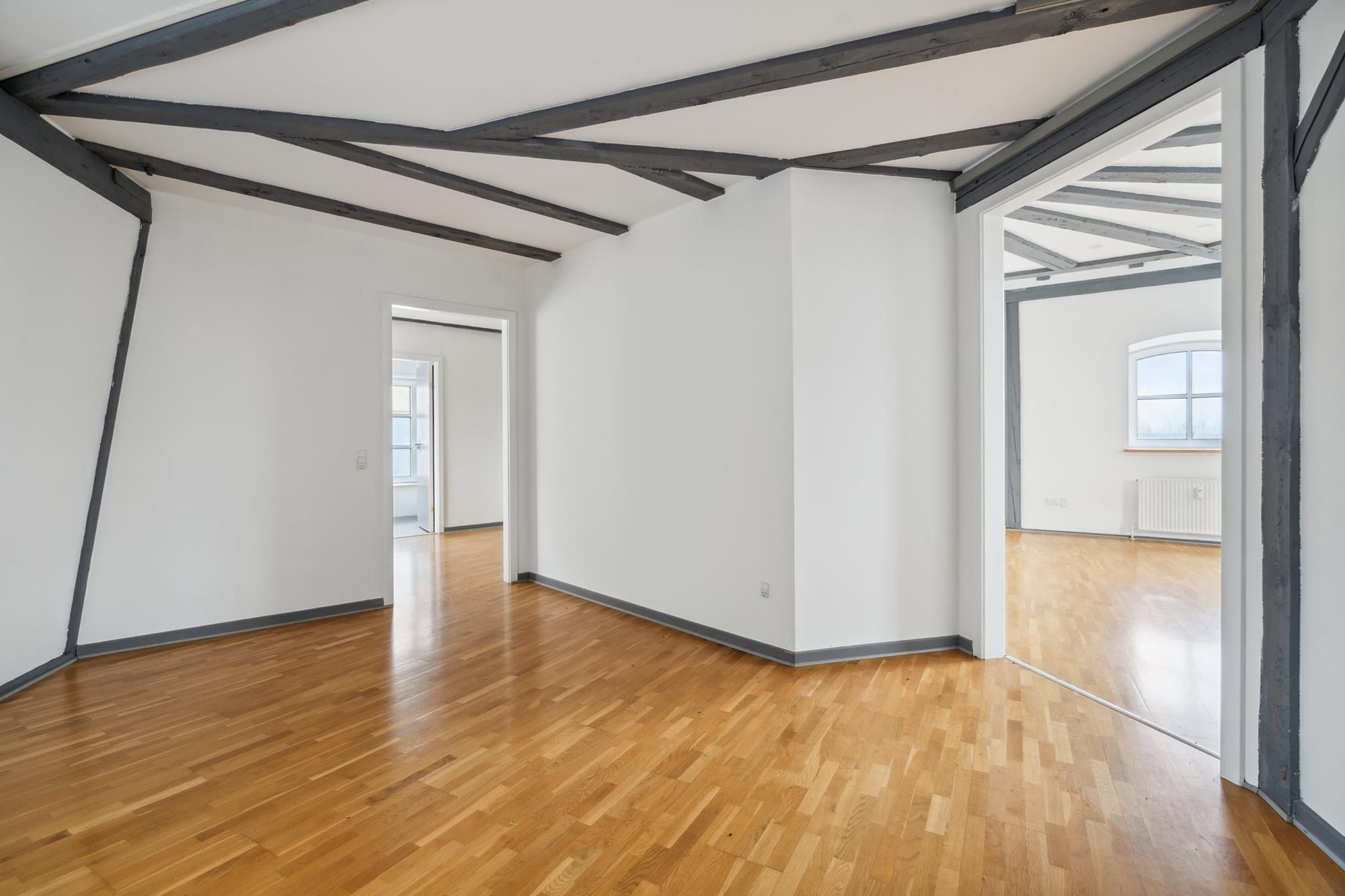 Stort hjørnerum med højloft, trægulv og hvide vægge samt to indgange til andre værelser og et vindue