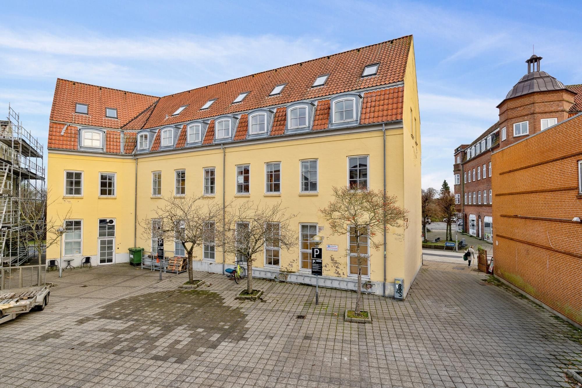 Gammel gule bygning med parkeringsskilt og 2 træer og 2 stolpe på forsiden.