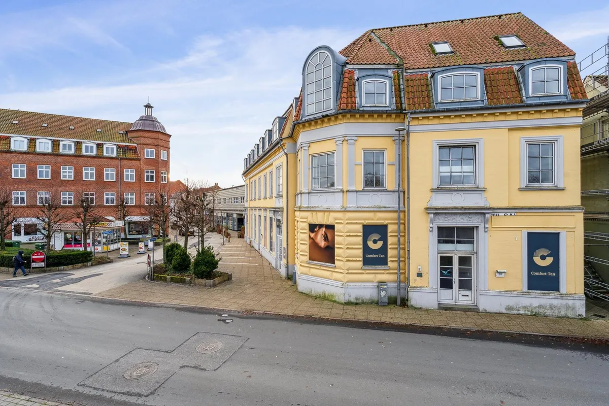 Bygning med facademaling og reklameskilte, mens en person går på gågaden. Butiksvinduer og søjler langs siderne.