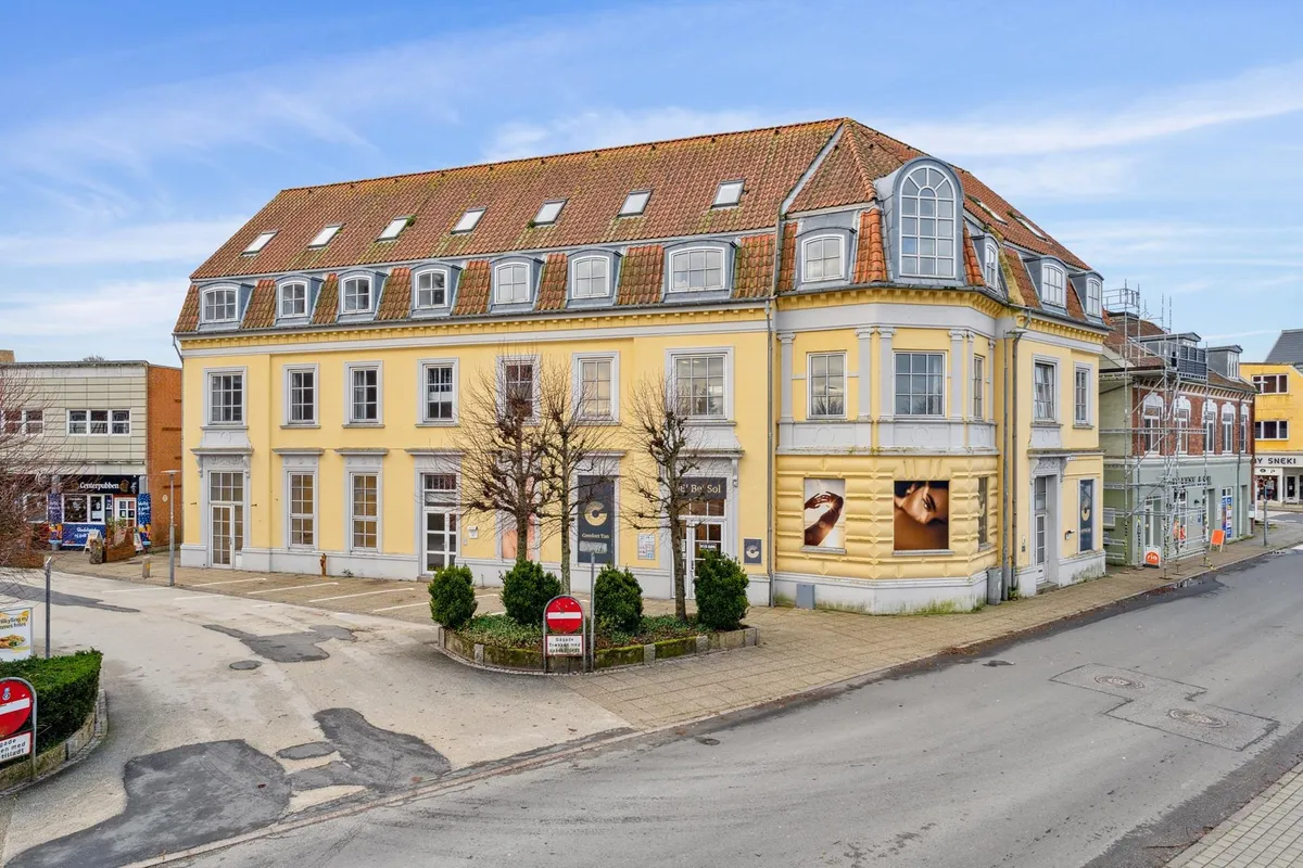 Gågade i Struer med grønne buske og et rødt skilt med en stribe, gult bygning med mange vinduer og et stort skilt med et