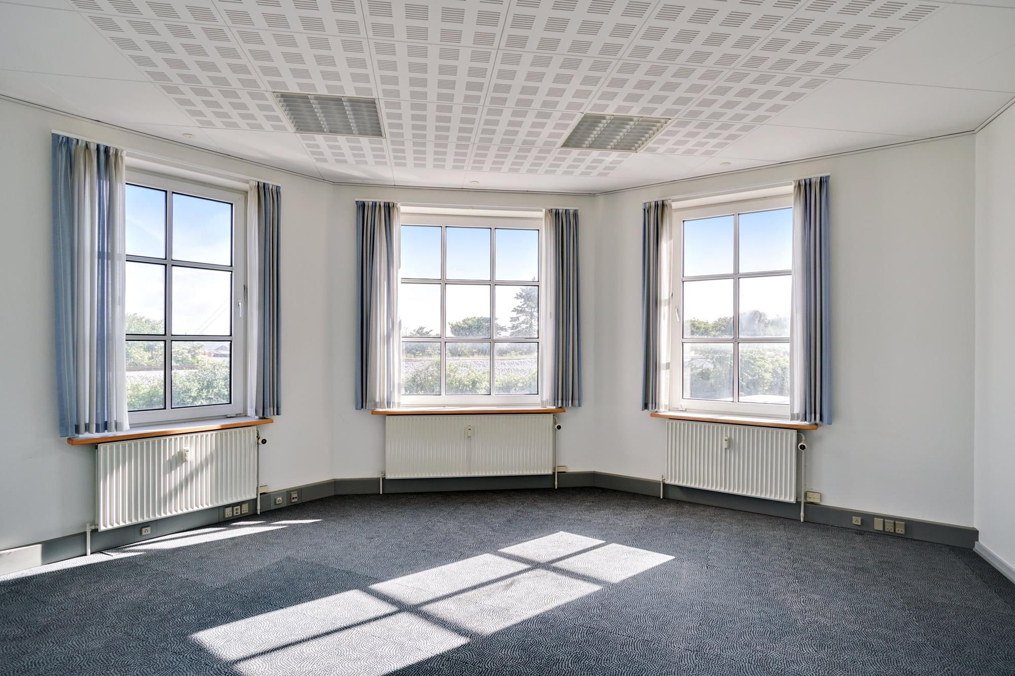 To vinduer med hvide gardiner og hvide radiatorer samt et loft med ventiler og lyse refleksioner på gulvet