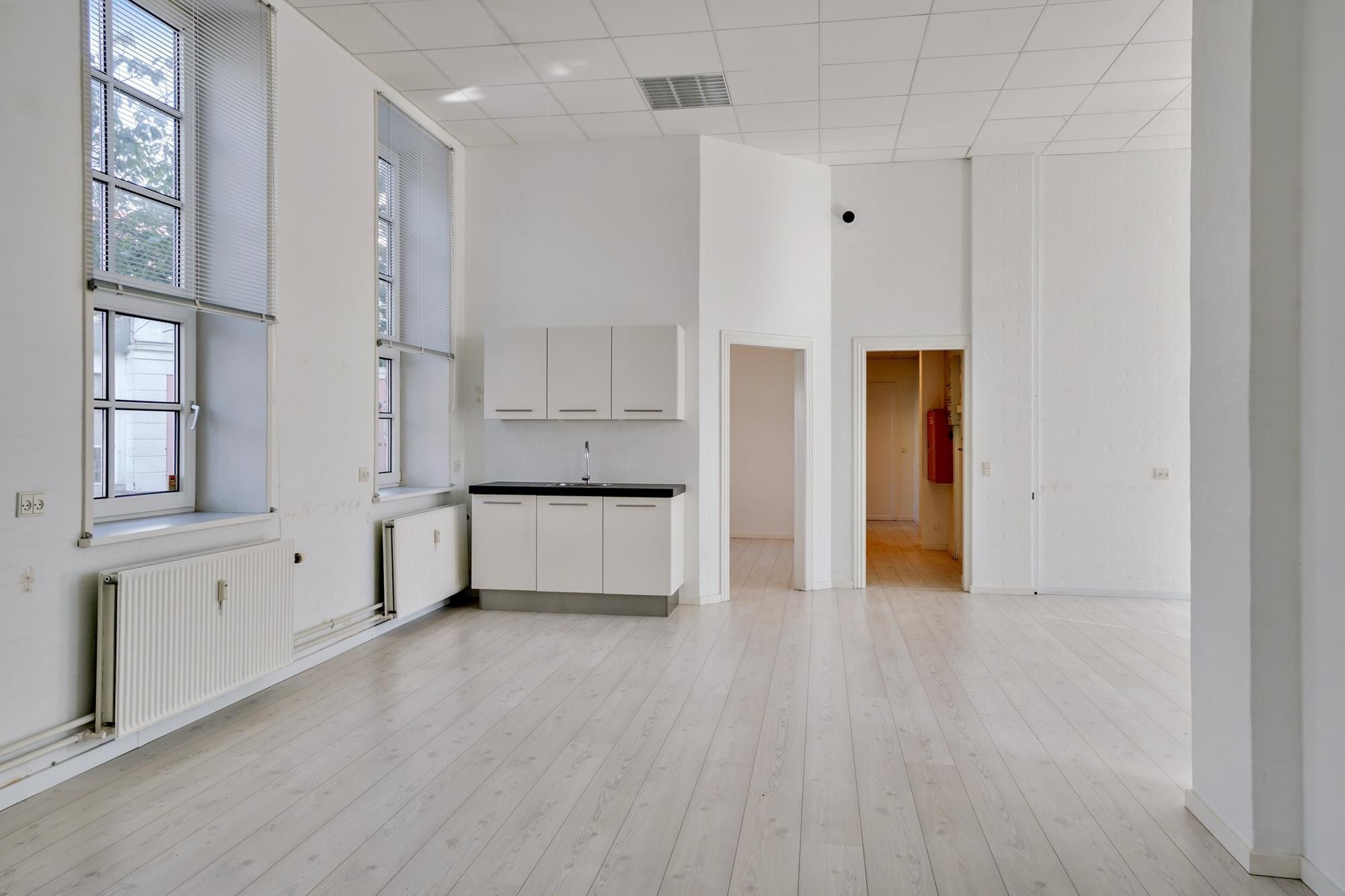 Minimalistisk kontorlokal med hvidt loft og hvide vægge. Køkken med sort bordplade og hvide skabsfronter. Vindue med