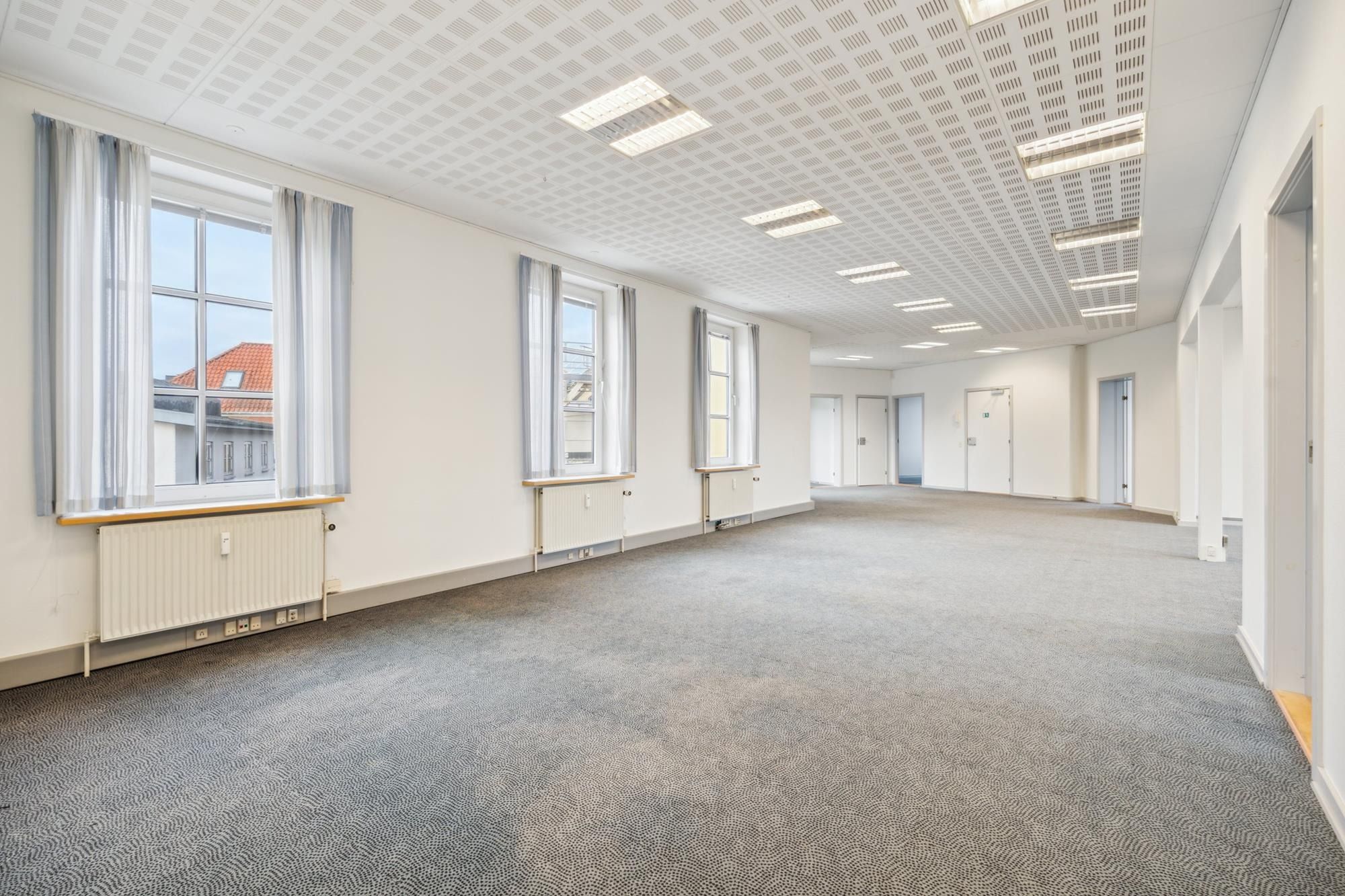 En stor, tom lejlighed med store vinduer, hvide vægge og lyse loftslamper. Der er en radiator under et af vinduerne og flere